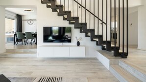 A modern living room with a staircase, a flat-screen TV mounted on a white console, and a dining area visible through an open door. - Villa Marita (Privlaka)