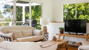 A living room with a sofa, a television on a wooden stand, and a grill outside the window. - The Treehouse Vincentia by Latitude South Coast (Vincentia)