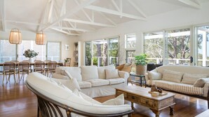 A spacious living room with a high ceiling, large windows, and a dining area with wicker furniture. - The Treehouse Vincentia by Latitude South Coast (Vincentia)