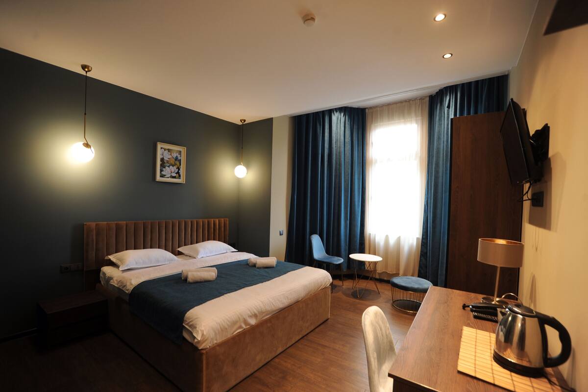 A hotel room with a bed, bedside table, chair, desk, lamp, and a television.