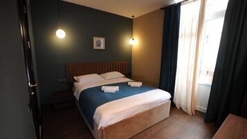 Ein ordentlich bezogenes Bett mit einer blau-weißen Decke, zwei Nachttischlampen, einem gerahmten Bild an der Wand und einem Fenster mit Vorhängen.