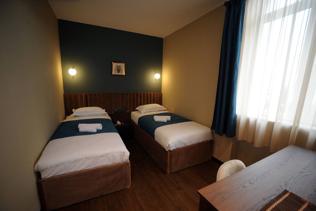 A hotel room with two single beds, a wooden headboard, a small table, and a window with curtains.