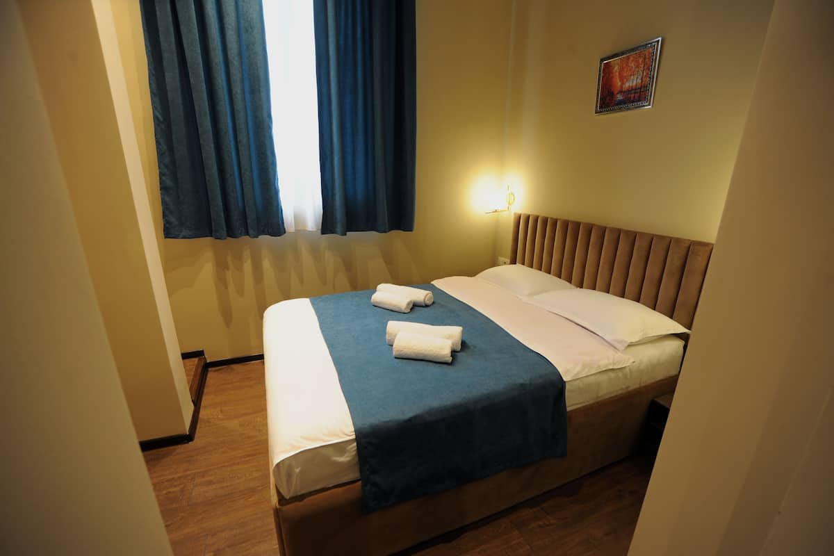 A neatly made bed with a blue bedspread and two rolled towels, a wall-mounted light, and a window with blue curtains.