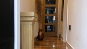 En hall med trägolv, en vit dörr, ett element och en vas med högt gräs.