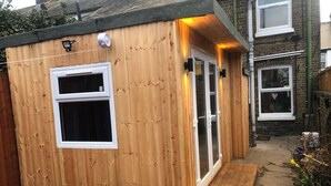 Ett litet trähus med ett enda fönster och en dörr, en liten träramp som leder fram till ingången, och ett hus av tegel i bakgrunden.