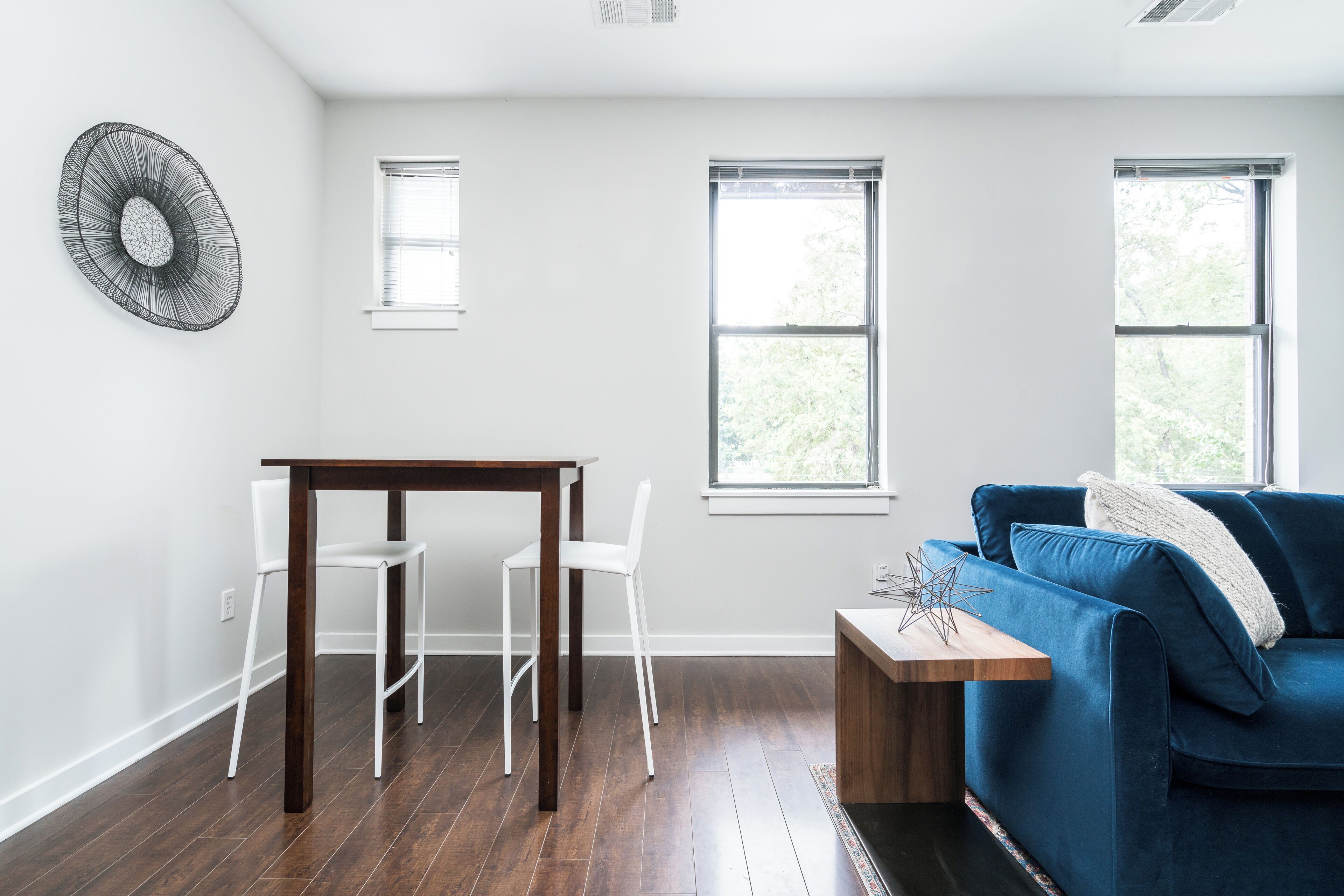 Una sala de estar moderna con una mesa de madera oscura, sillas blancas, un sofá azul y dos ventanas con persianas.