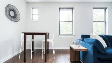 Una sala de estar moderna con una mesa de madera oscura, sillas blancas, un sofá azul y dos ventanas con persianas.