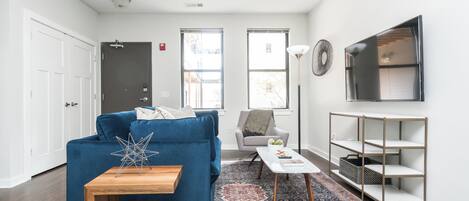 Una sala moderna con un sofá azul, una pequeña mesa de madera, un tapete estampado, un sillón gris y un televisor montado en la pared.