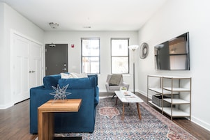 A modern living room with a blue sofa, a small wooden table, a patterned rug, a grey armchair, and a TV mounted on the wall.