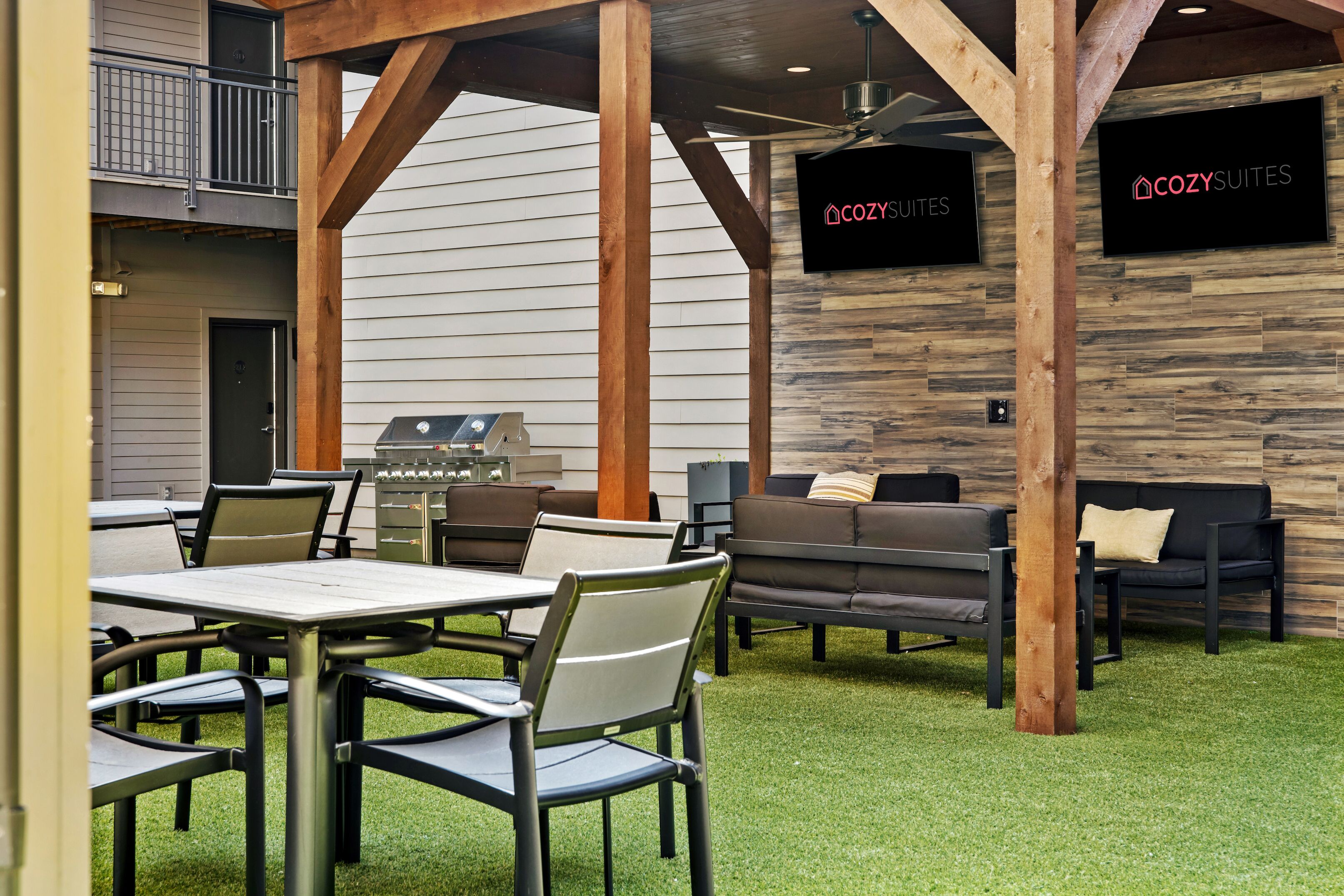 Un patio exterior con una parrilla, asientos y una pantalla de televisión mostrando 'COZY SUITES'.