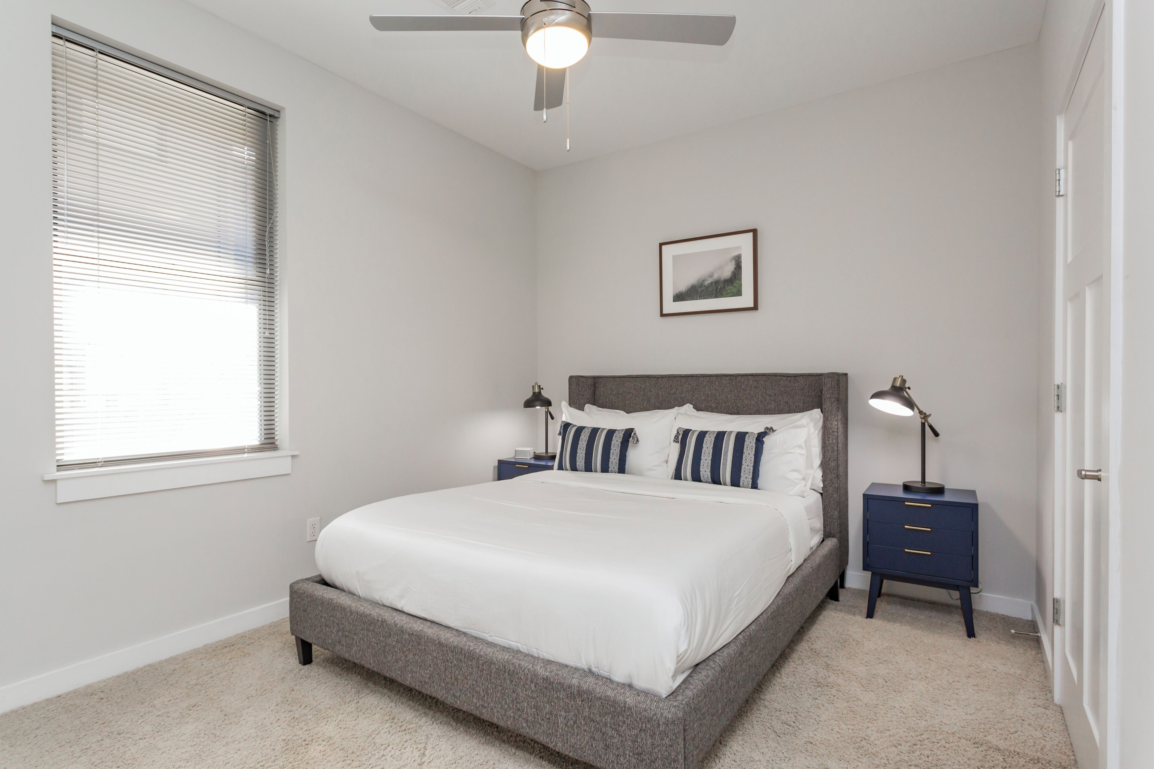 A bedroom with a bed, a ceiling fan, a nightstand, a bedside lamp, a framed picture on the wall, and a window with blinds.