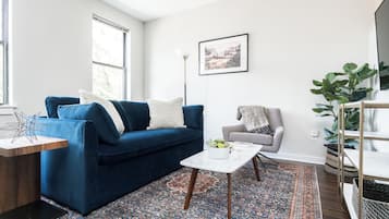 Una sala de estar con un sofá azul, una mesa de centro blanca, un tapete estampado, un sillón gris y un ventanal grande.
