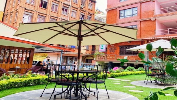 An outdoor patio area with a table and chairs under a large umbrella, surrounded by greenery and a brick building in the background. - MeroStay 195  Hotel Garden  Pvt Ltd (Kathmandu)