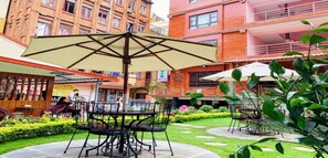 An outdoor patio area with a table and chairs under a large umbrella, surrounded by greenery and a brick building in the background. - MeroStay 195  Hotel Garden  Pvt Ltd (Kathmandu)