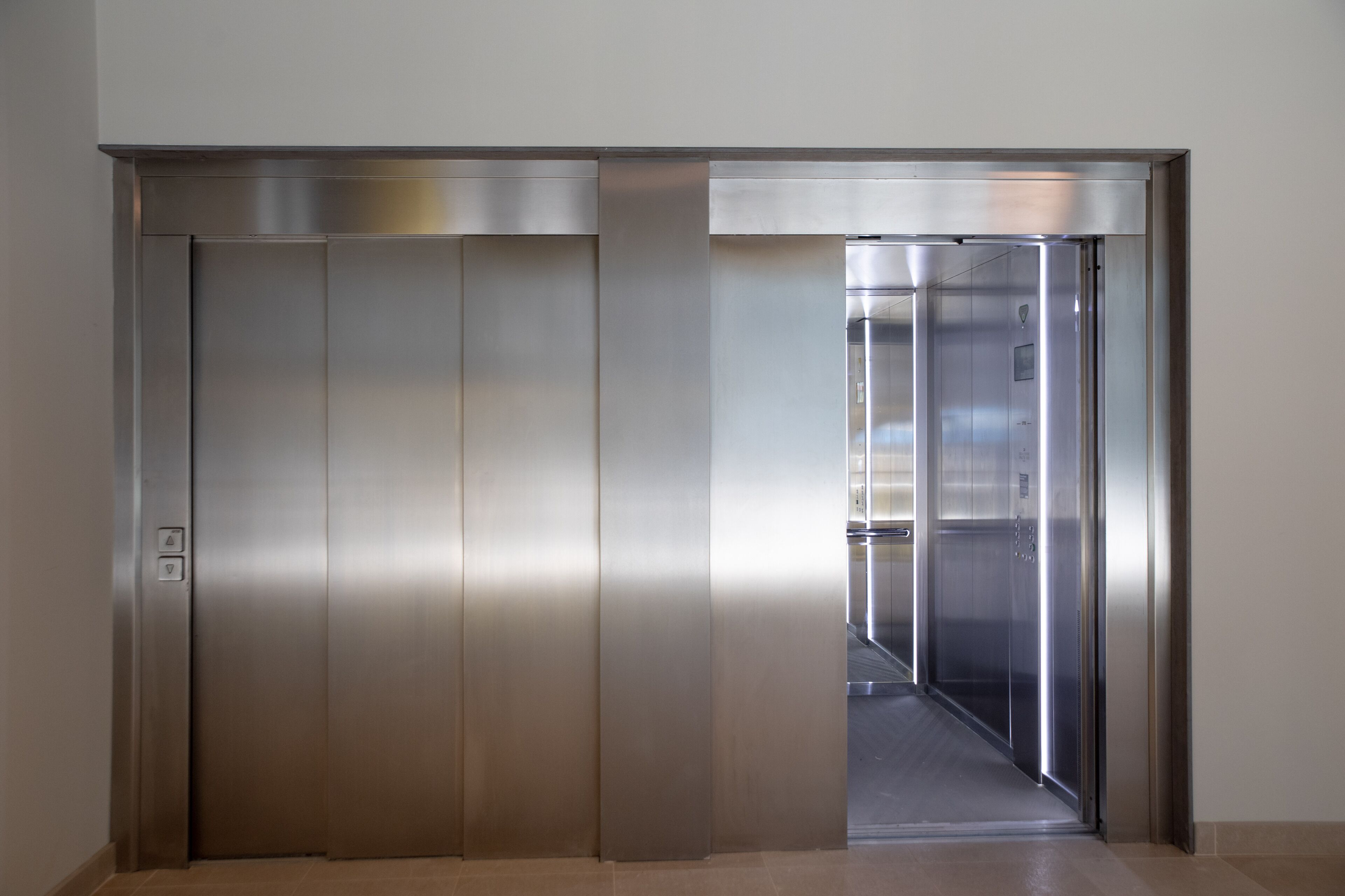 A metallic elevator with a modern design, featuring a control panel on the left and a clear glass door.