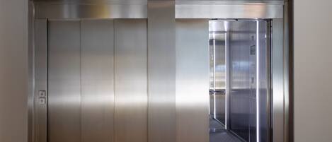 Un ascensor metálico de diseño moderno, con un panel de control a la izquierda y una puerta de vidrio transparente.