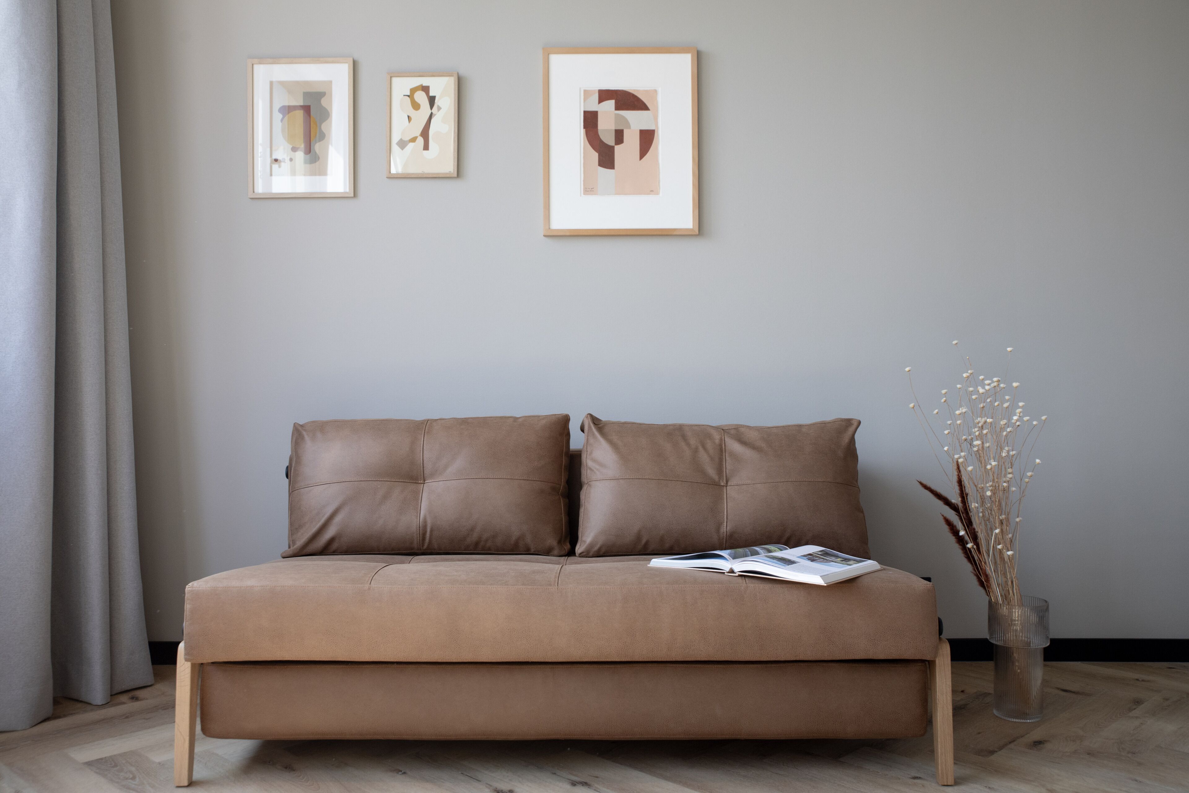 A neutral-toned room with a brown sofa, two framed artworks on the wall, and a vase with dried plants on the floor.
