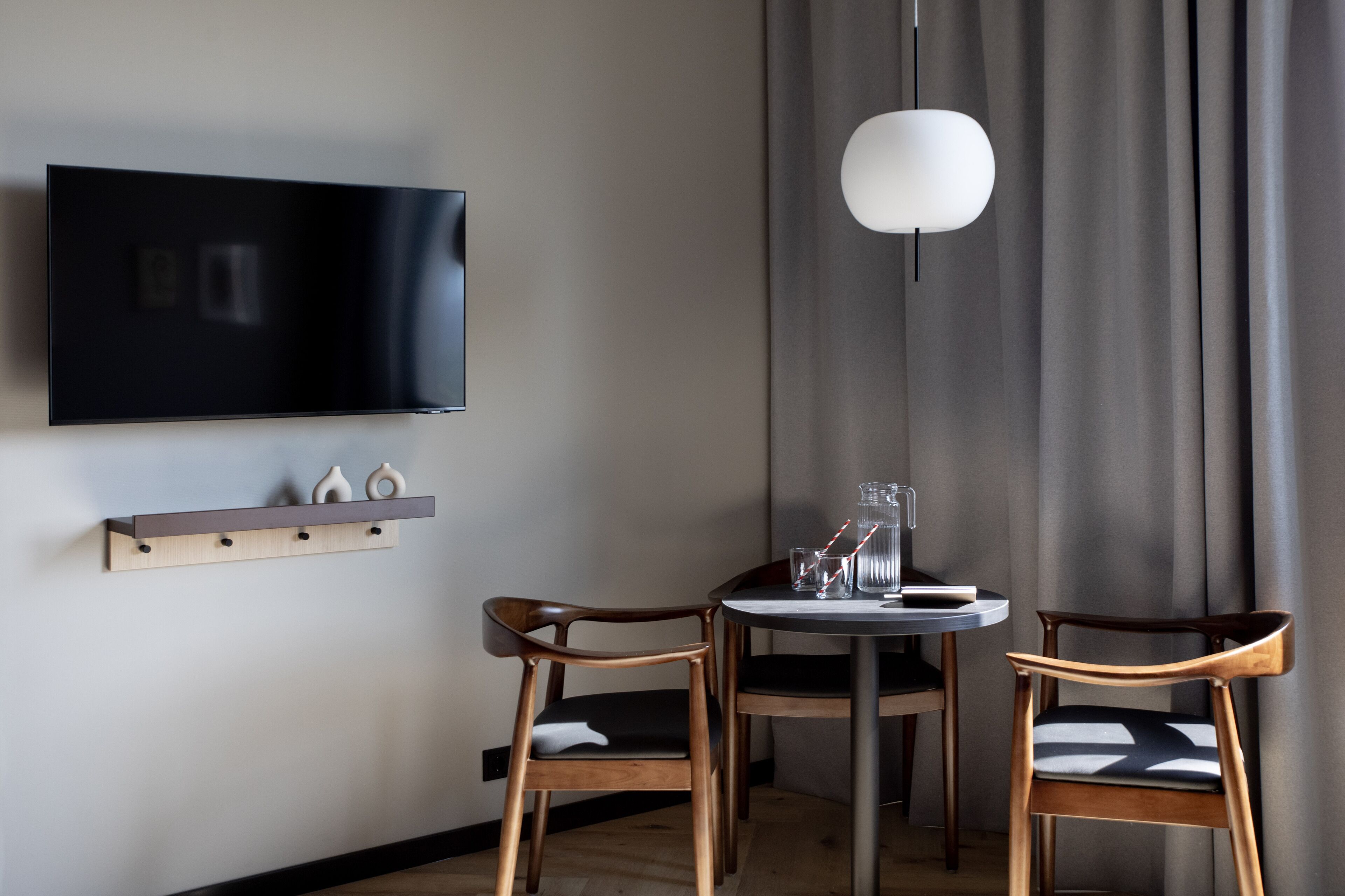 Quarto moderno com televisão de tela plana na parede, uma mesa redonda com duas cadeiras de madeira e uma luminária pendente.