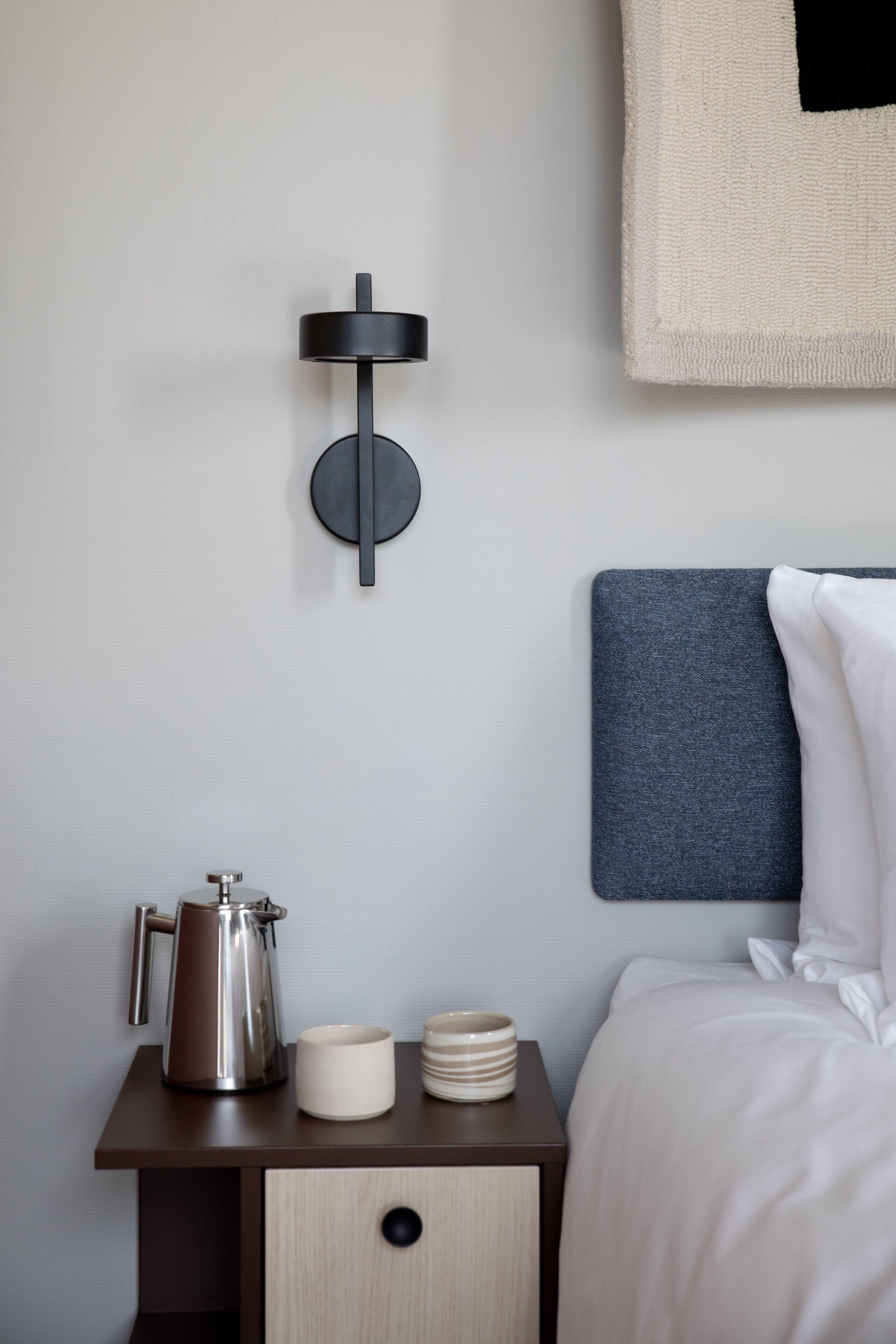 A neatly arranged hotel room with a bed, a bedside table with a kettle, cups, and a small cabinet.