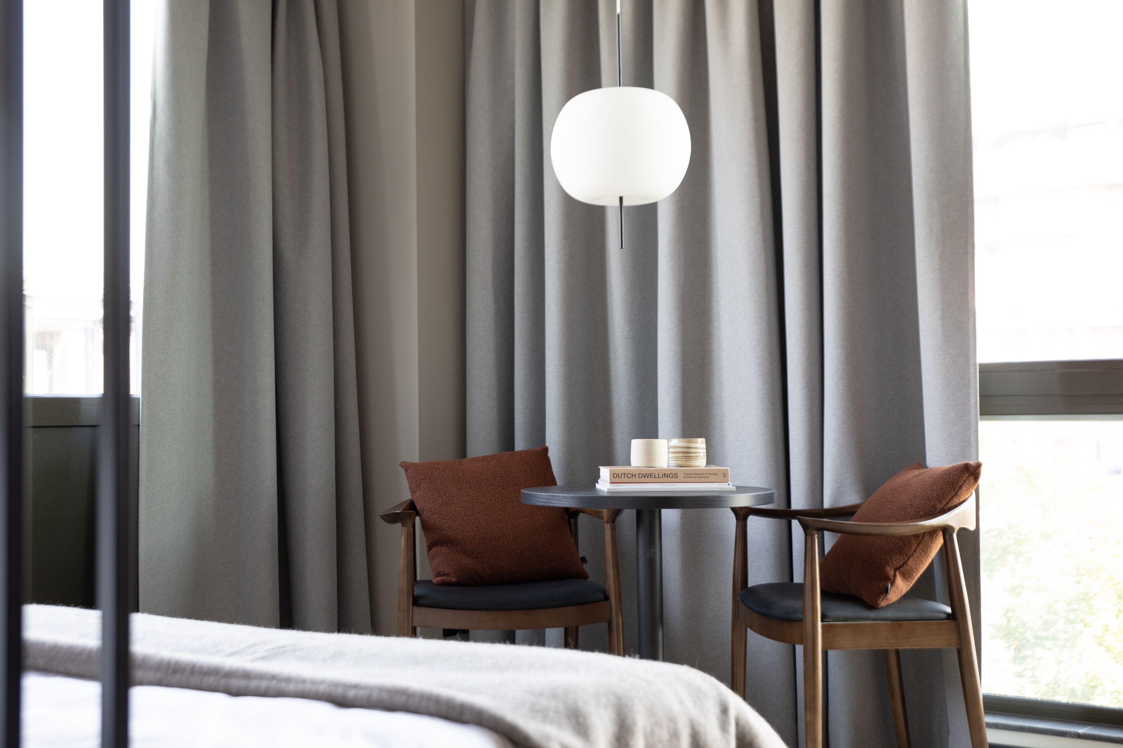 Une chambre d’hôtel avec un lit, deux chaises, une petite table avec un livre et des bougies, et une grande fenêtre avec des rideaux.