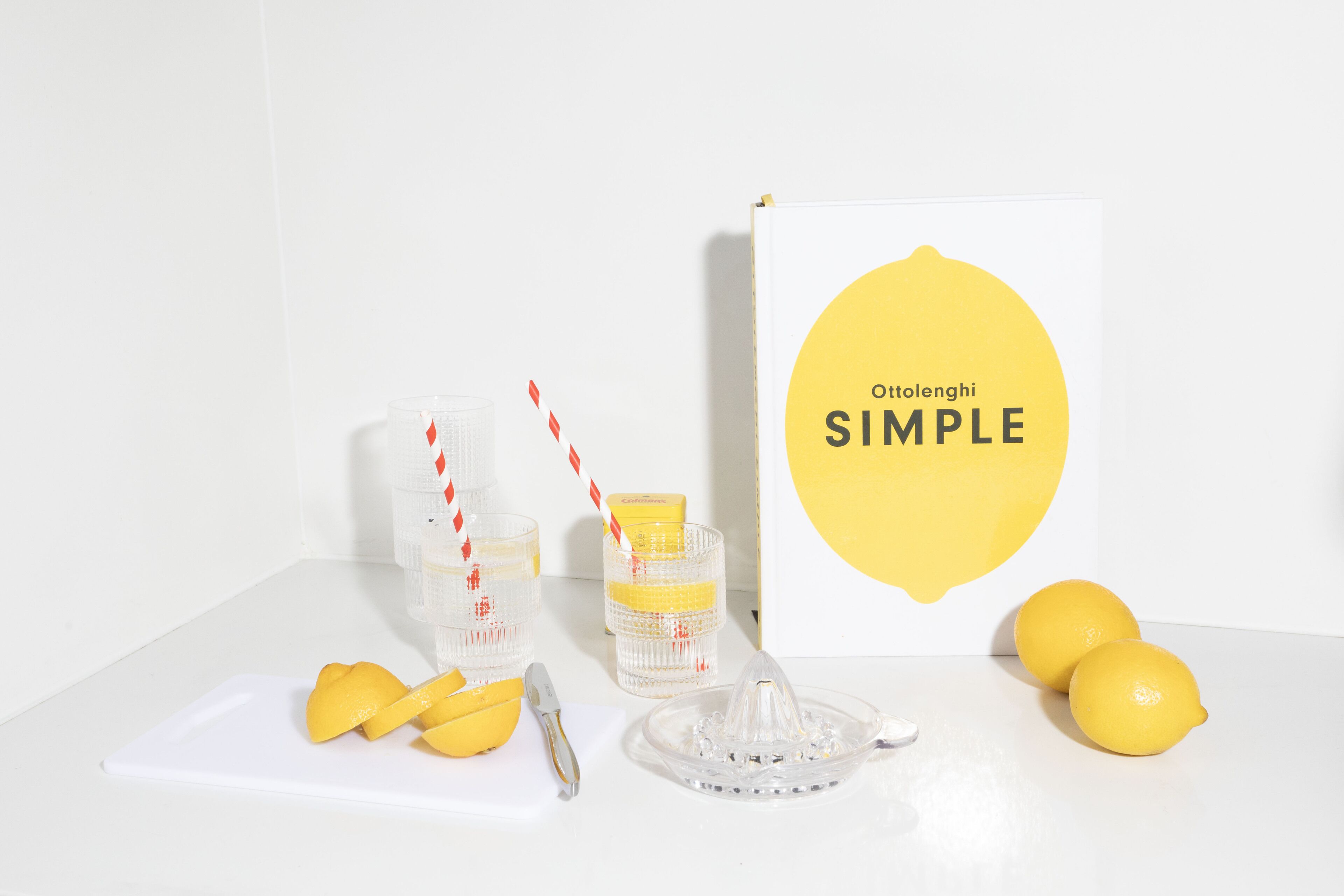 Una mesa blanca con un exprimidor de limones, dos limones, un cuchillo y un vaso con pajita.
