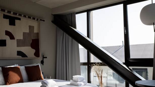 A hotel room with a large bed, a view of the city through large windows, a modern light fixture, and a decorative wall piece. - Bob W Amsterdam Noord (Amsterdam)