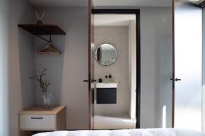 Un dormitorio moderno con cama, mesita de noche con un jarrón, estante colgante, armario y un baño visible a través de una puerta abierta.