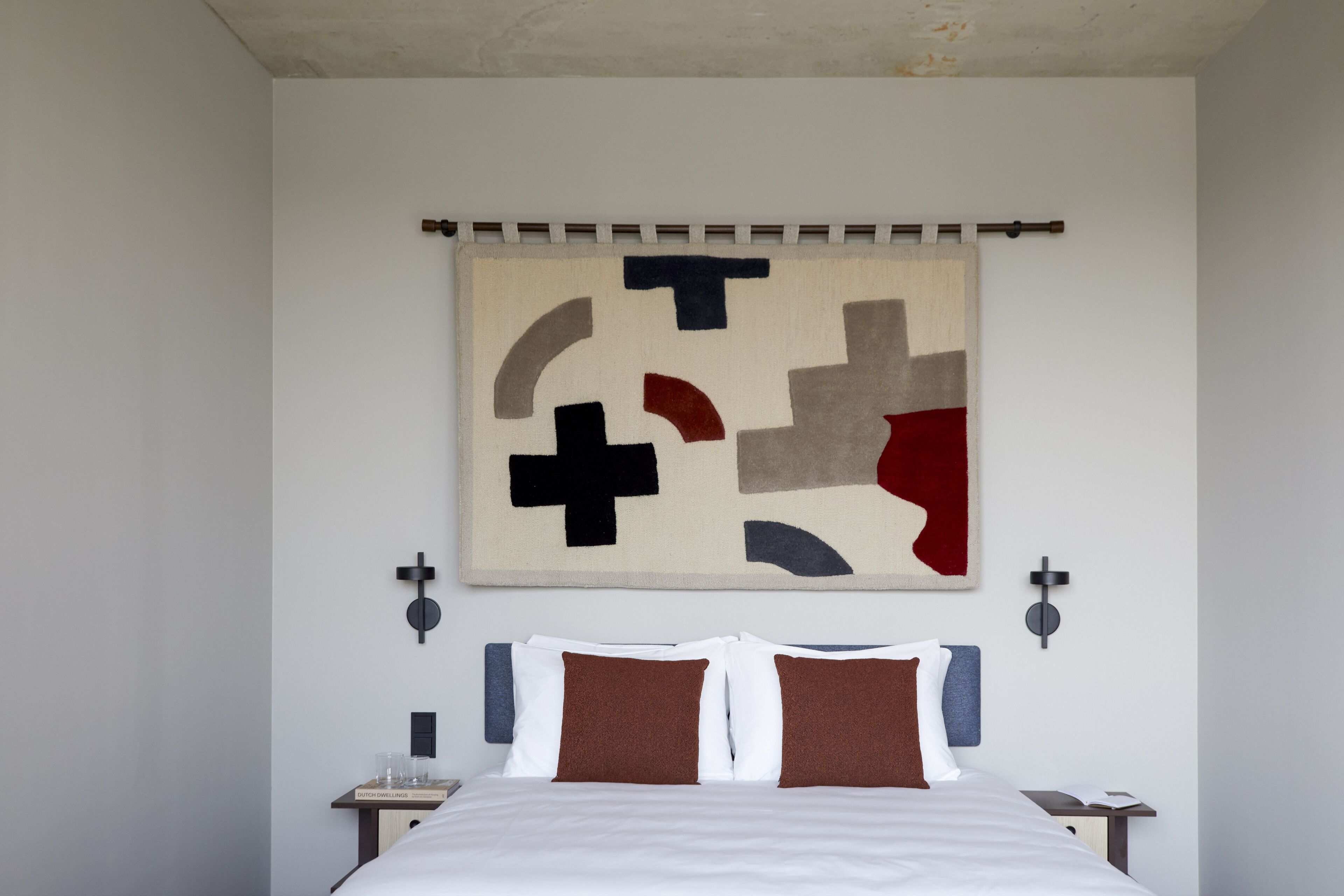 A modern hotel room with a large bed, a wall-mounted abstract artwork, and a simple bedside table with a lamp.