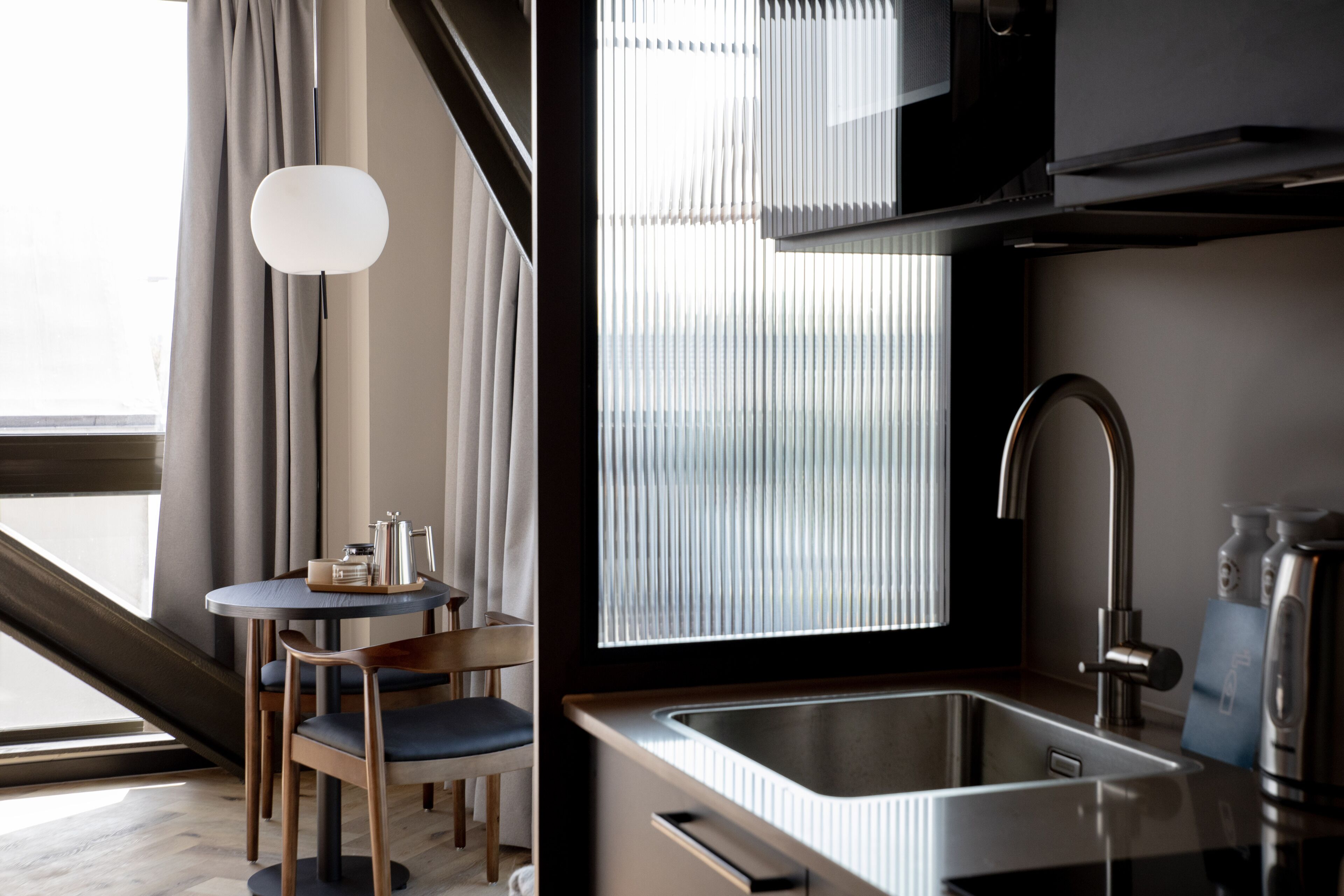 A modern kitchen with a stainless steel sink, a round dining table with chairs, and a large window with blinds.