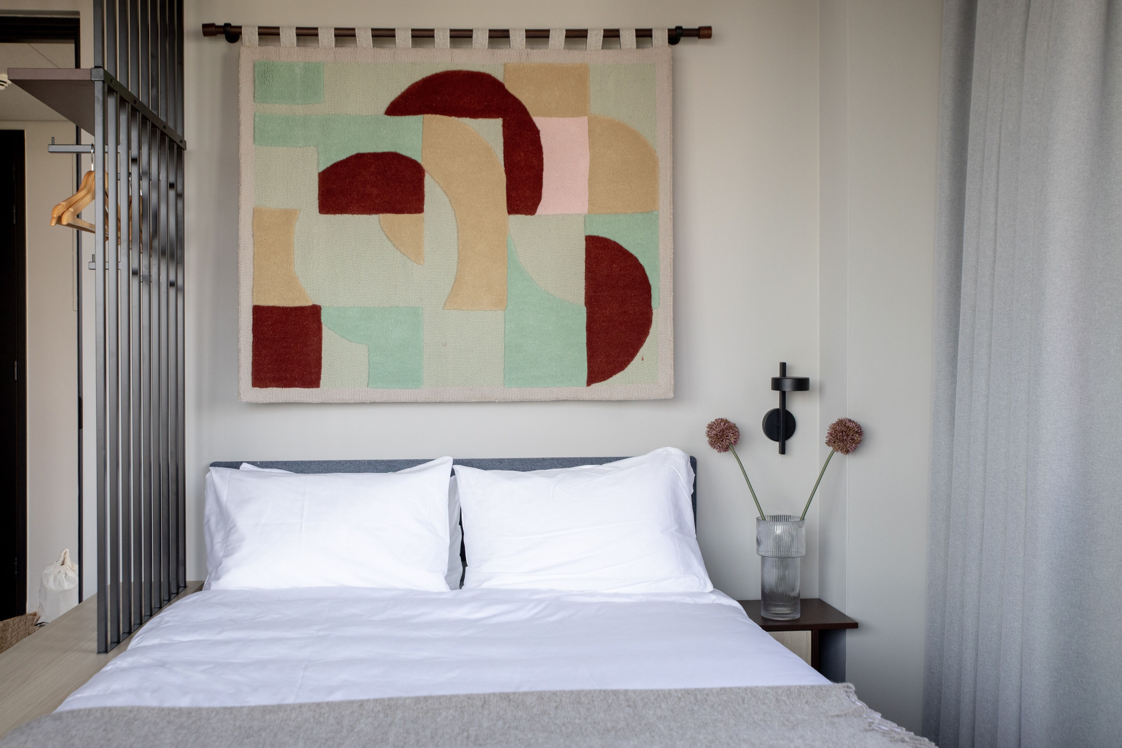 Un dormitorio moderno con una cama grande, un cuadro abstracto geométrico, una mesita con un florero y una cortina.