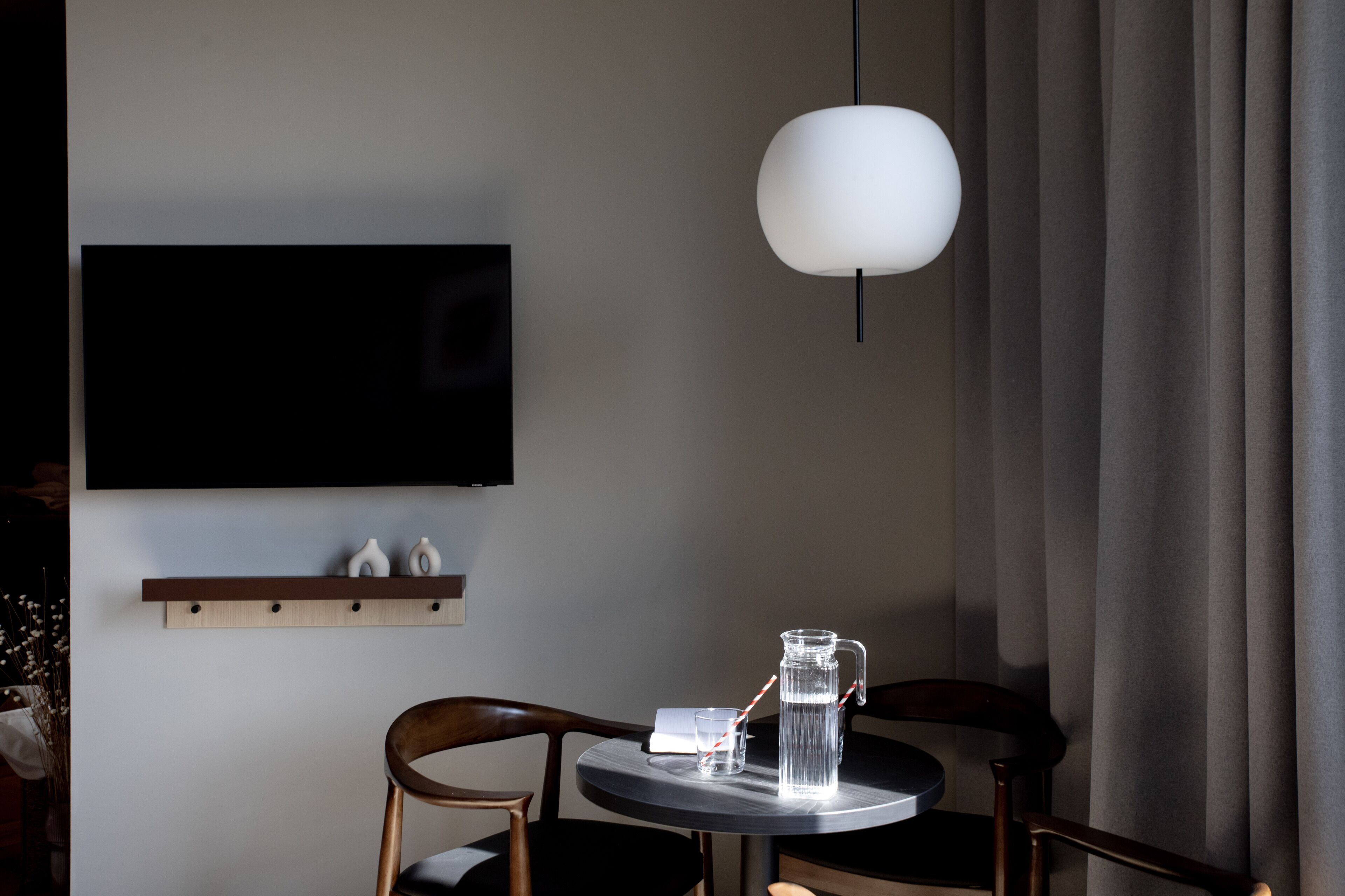 A modern room with a round table, a mounted TV, a hanging light fixture, and a window with curtains.