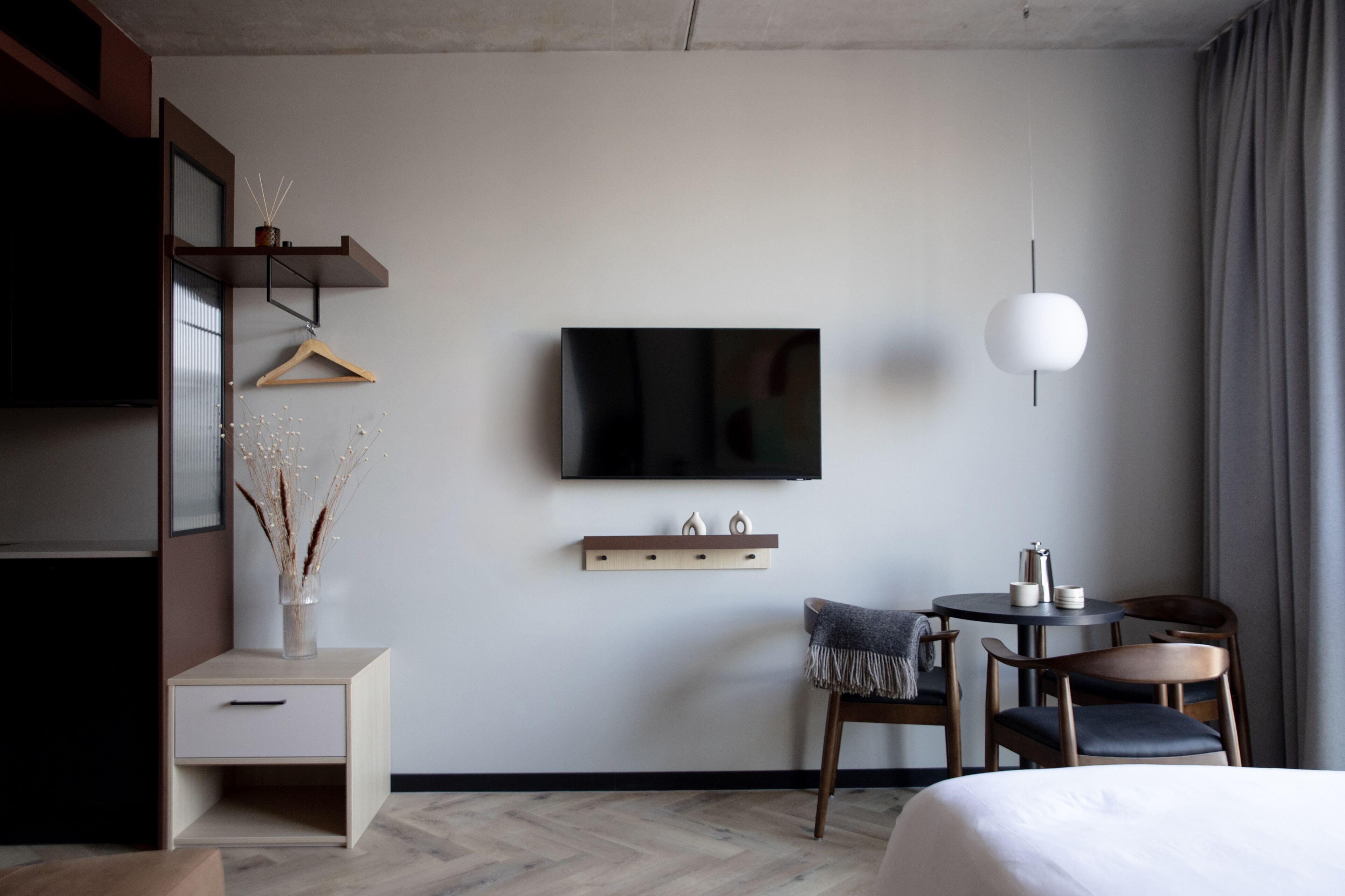 A modern hotel room with a bed, a desk with a chair, a TV mounted on the wall, and a hanging light fixture.