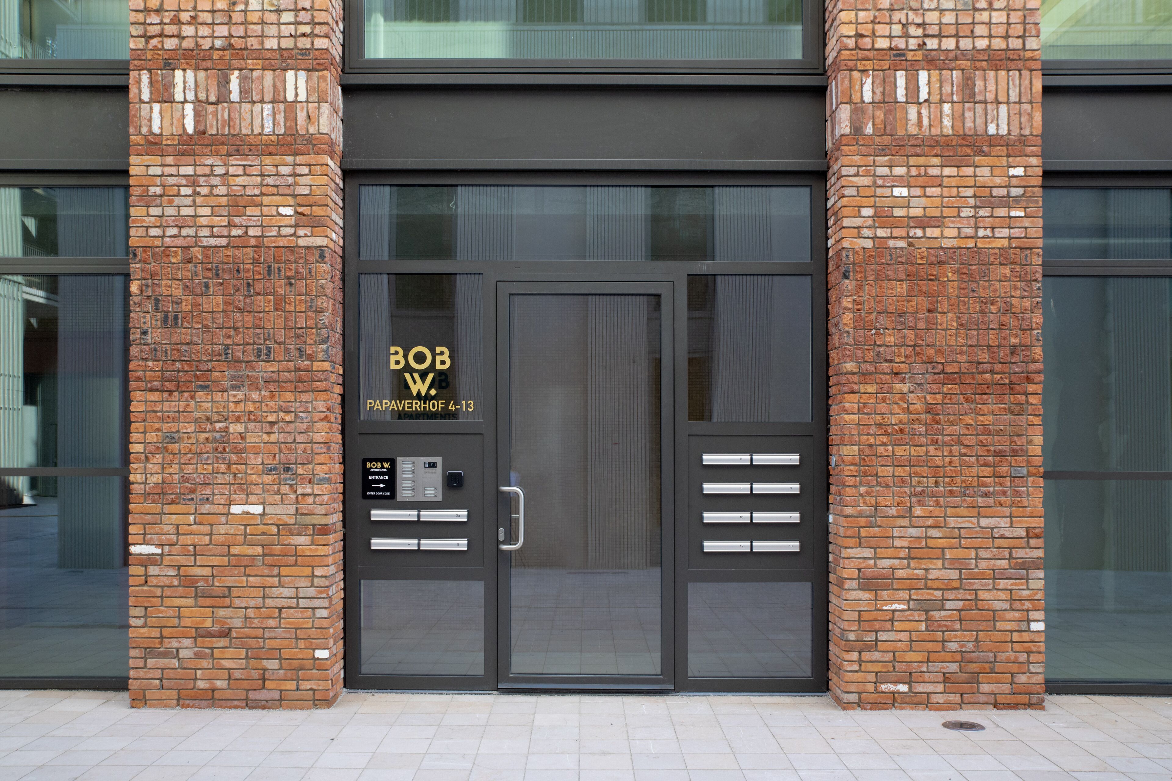 A modern building entrance with a glass door, a brick facade, and a sign indicating "BOB W PAPAVERHOF 4-13".