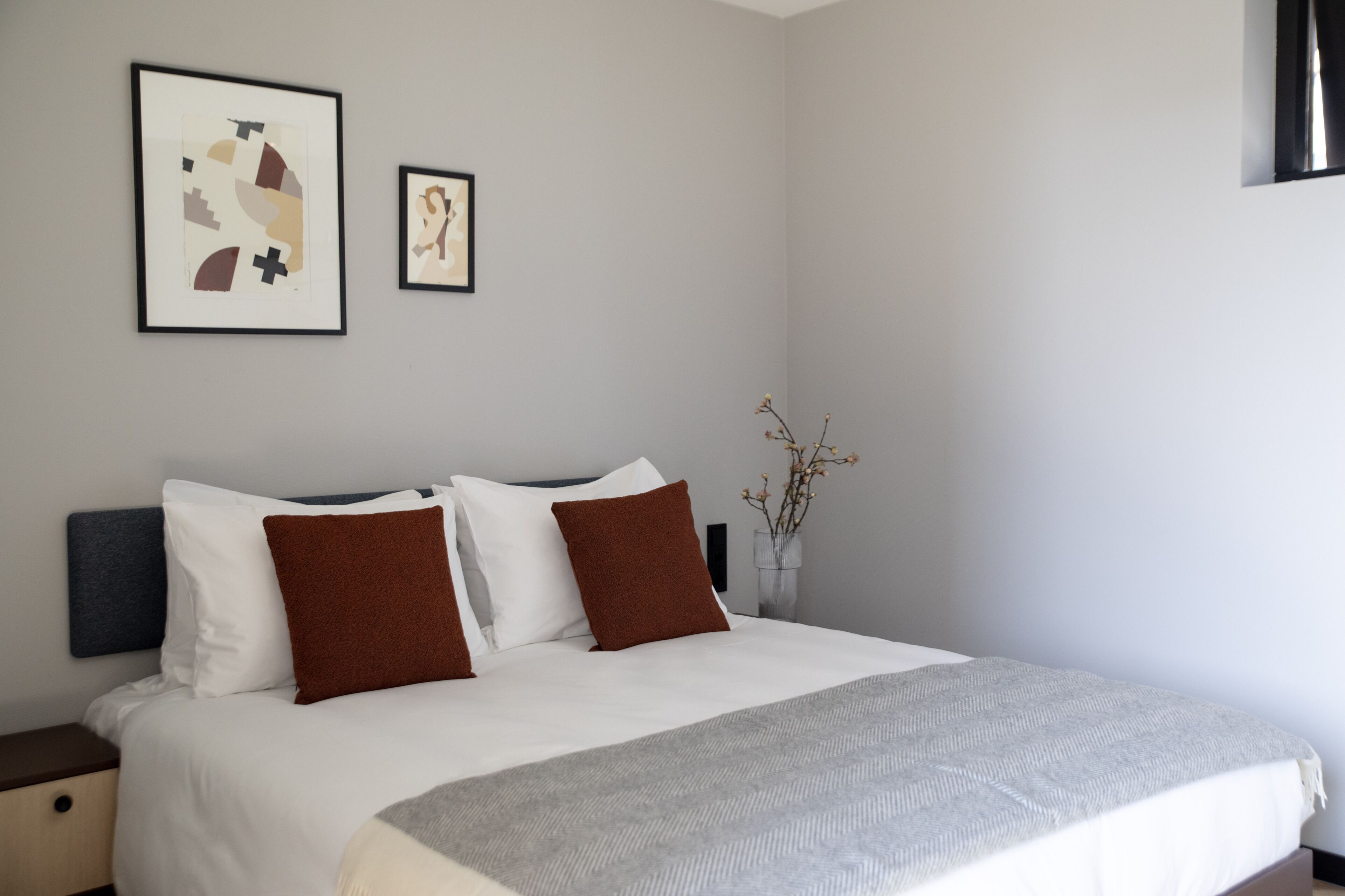 A neatly made bed with white and brown bedding, a gray throw blanket, and two framed artworks on the wall.