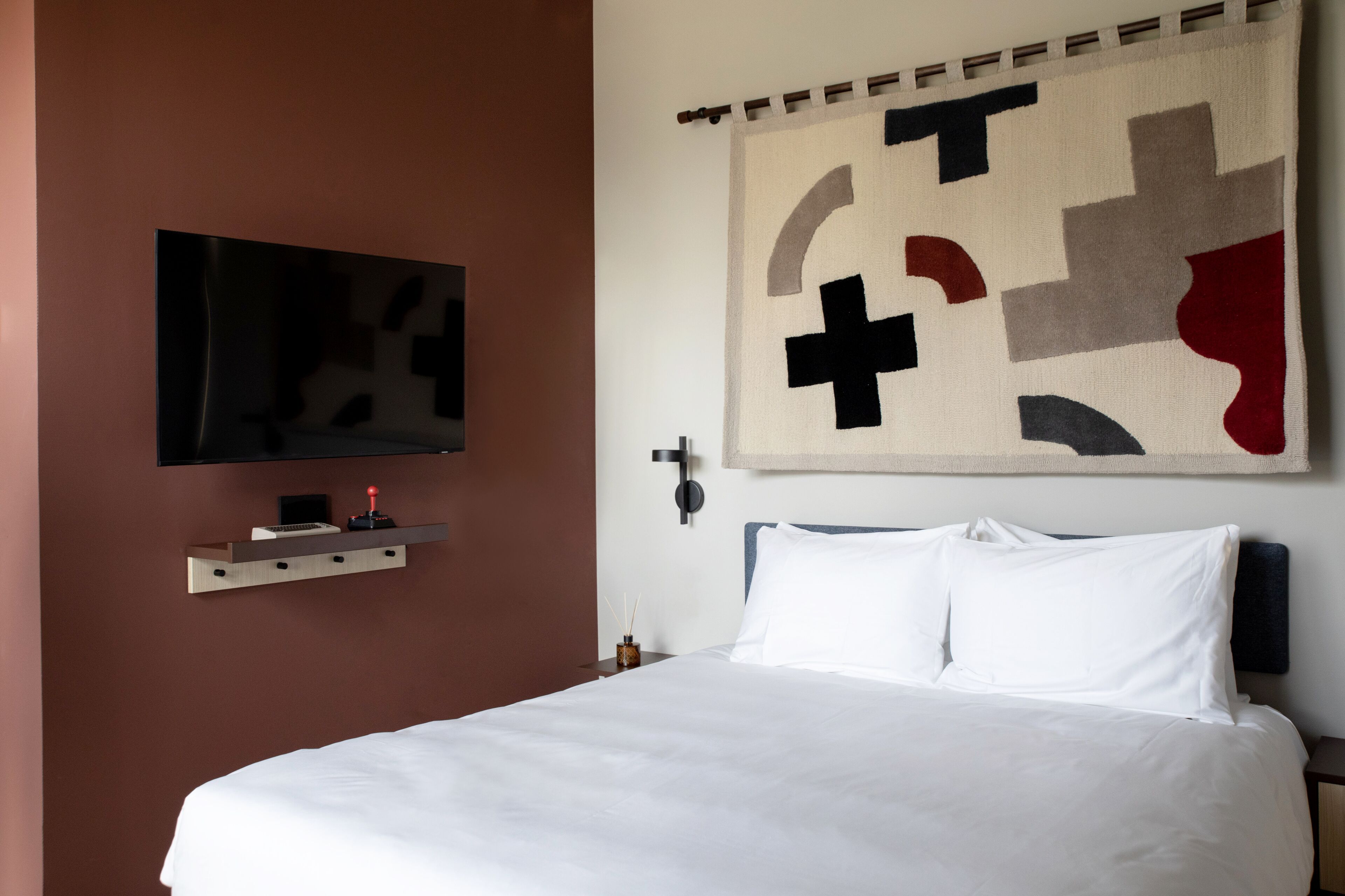 A hotel room with a bed, a mounted TV, a wall-mounted shelf, and a large abstract artwork.