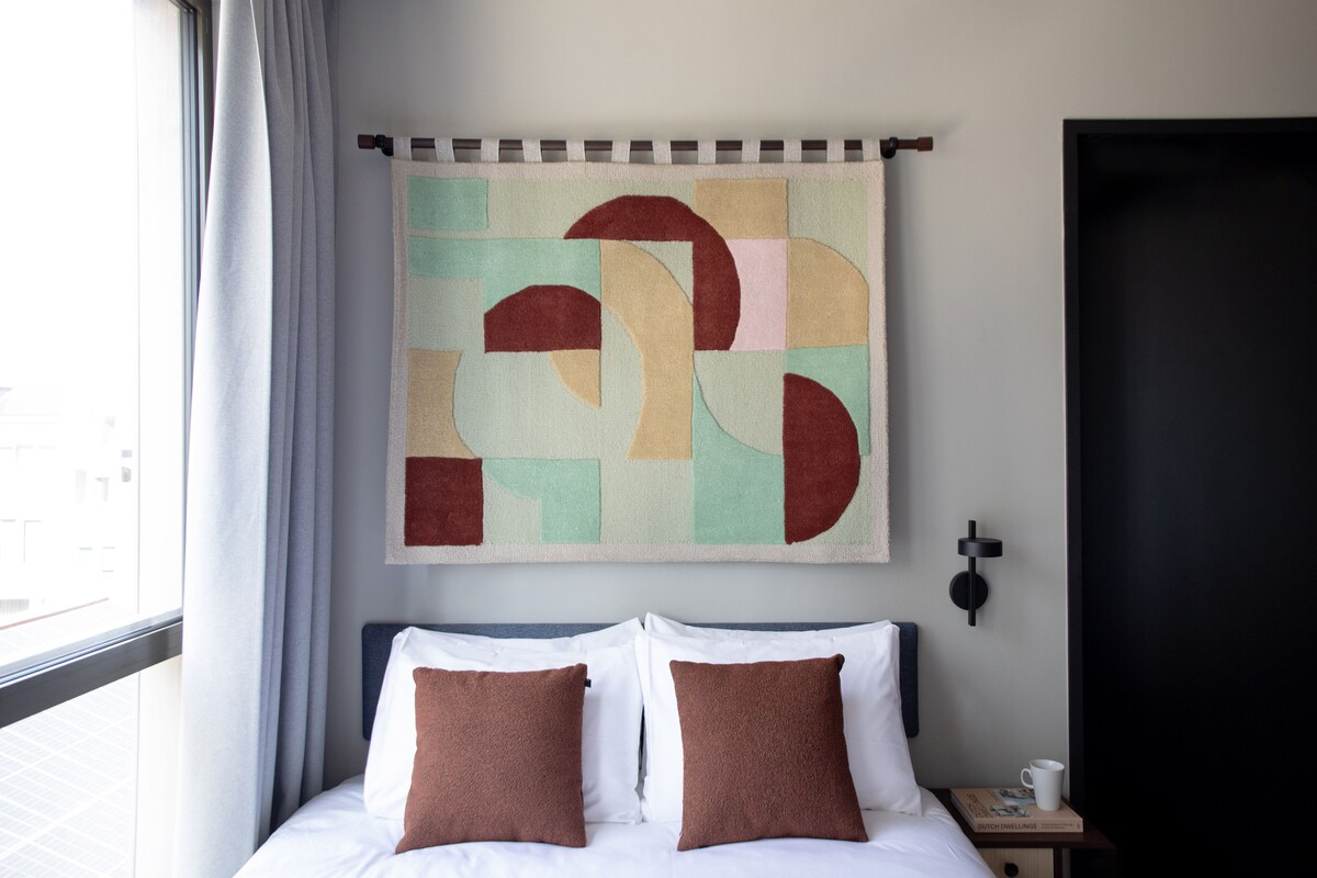 A neatly made bed with two pillows, a wall-mounted abstract artwork, a small bedside table with a book and a cup, and a window with curtains.