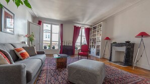 En stue med en grå sofa, en rød lænestol, et mønstret tæppe, en reol og en pejs.