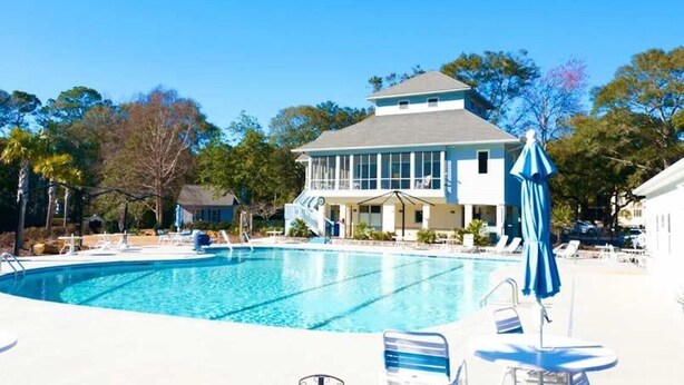 Une piscine entourée d’arbres, avec des chaises longues et des parasols, et un bâtiment doté d’un balcon.
