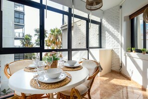 A dining area with a round table set for four, surrounded by wicker chairs, with a view of palm trees and a building through large windows. - 0. Valdecantos Suites (Marbella)