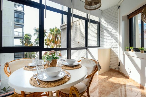 Un coin repas avec une table ronde dressée pour quatre personnes, entourée de chaises en osier, et offrant une vue sur des palmiers et un bâtiment à travers de grandes fenêtres.