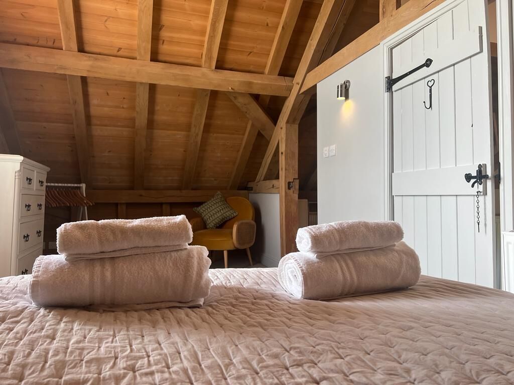 A bedroom with a wooden ceiling, a bed with a quilted bedspread, two rolled towels, a white wardrobe, and a door with a heart-shaped handle.