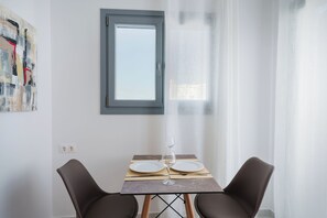 A dining area with a table set for two, a window, and a painting on the wall. - Bedspot Apartments Paros (Paros)