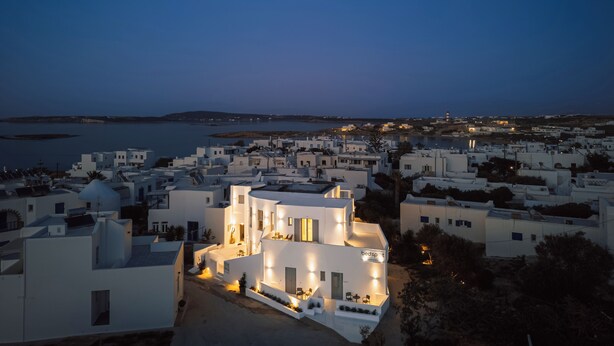A coastal town at dusk with white buildings, a prominent building with a sign 'bedsp' indicating a hotel, and a view of the sea and distant hills. - Bedspot Apartments Paros (Paros)