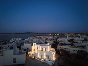 Un pueblo costero al anochecer con edificios blancos, un edificio destacado con el letrero 'bedsp' que indica un hotel, y vistas al mar y colinas distantes.