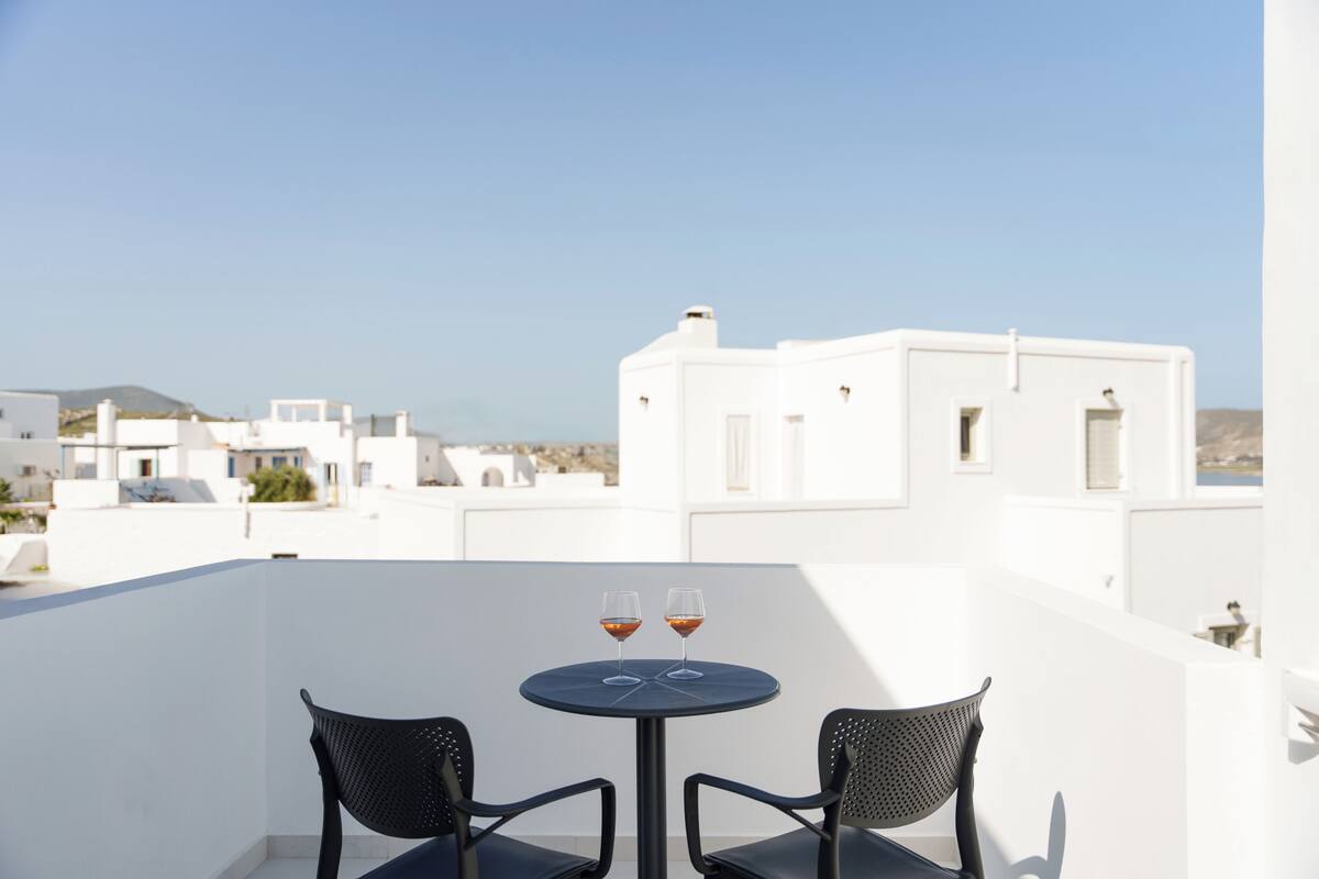 A balcony with two black chairs and a small round table, two glasses of red wine, and a view of white buildings and a mountain in the background.