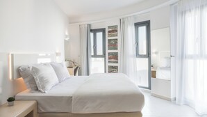 A modern bedroom with a large bed, a bedside table with a lamp, a small plant, and a view of the city through large windows. - Bedspot Apartments Paros (Paros)