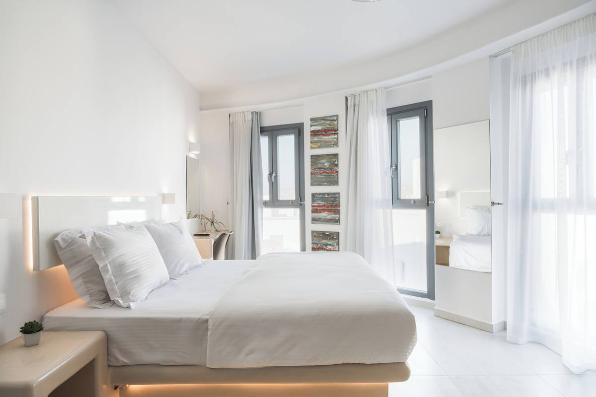 A modern bedroom with a large bed, a bedside table with a lamp, a small plant, and a view of the city through large windows.