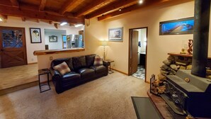 A cozy living room with a fireplace, a sofa, and a kitchen area visible through an open door. - Juneau Cabin near Eaglecrest & Trails (Juneau)