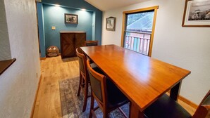 A dining area with a wooden table and chairs, a blue accent wall, a window with a view, and a door leading to another room. - Juneau Cabin near Eaglecrest & Trails (Juneau)
