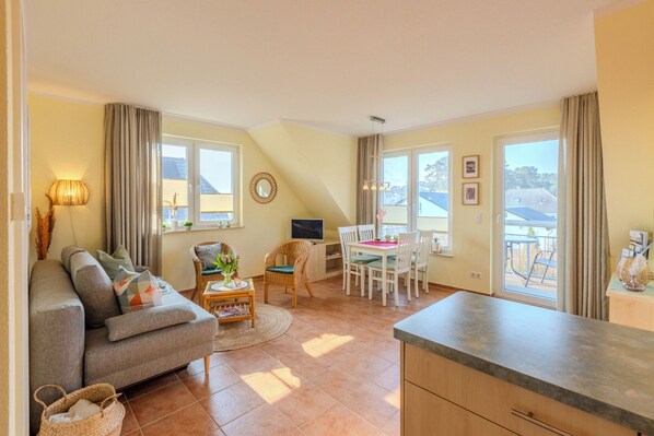 A cozy living room with a sofa, a dining table, and a kitchen area. There are windows with curtains, a TV, and a small plant on the windowsill.